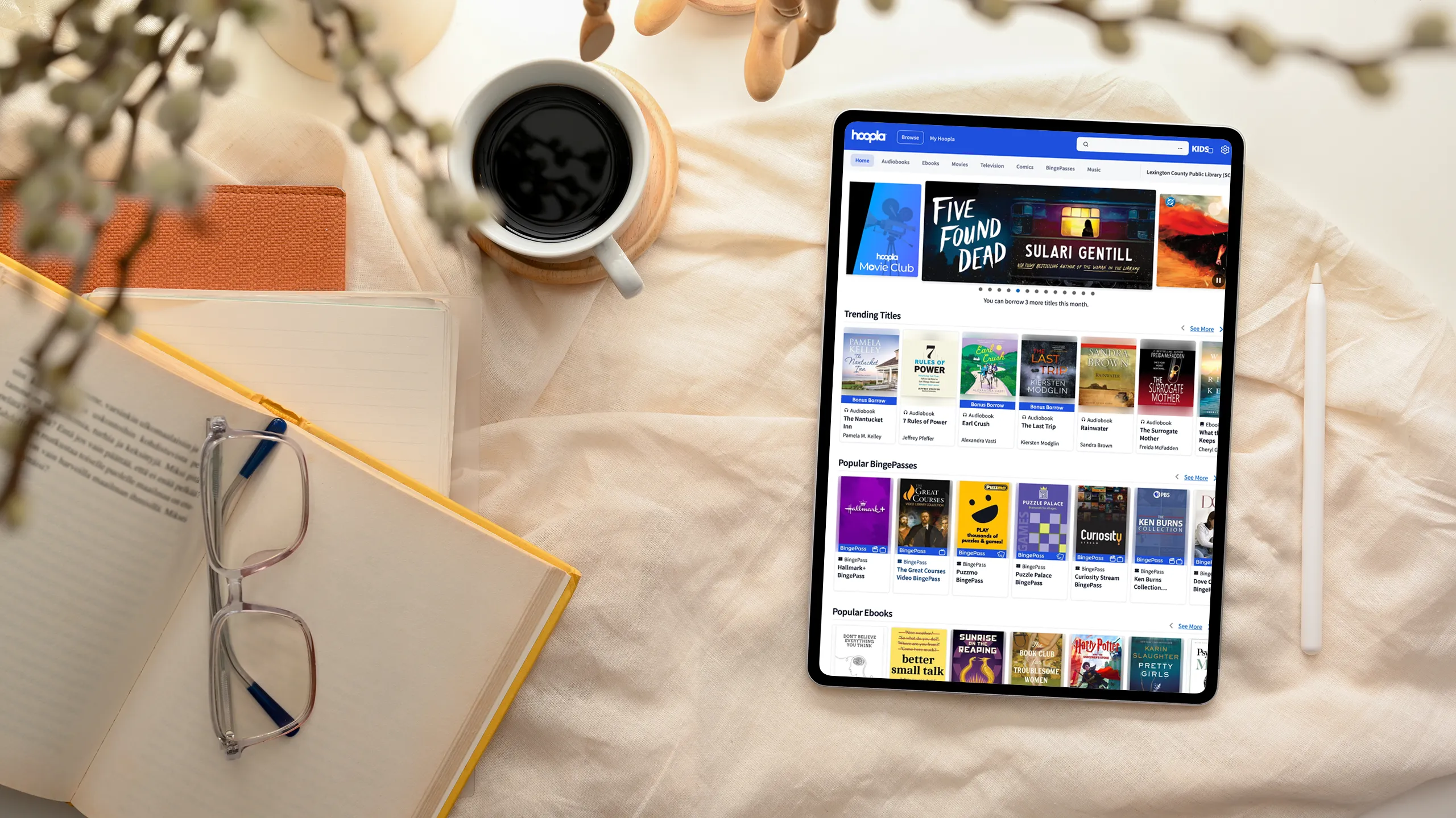 Tablet displaying the Hoopla website next to a book, glasses, notebook and coffee