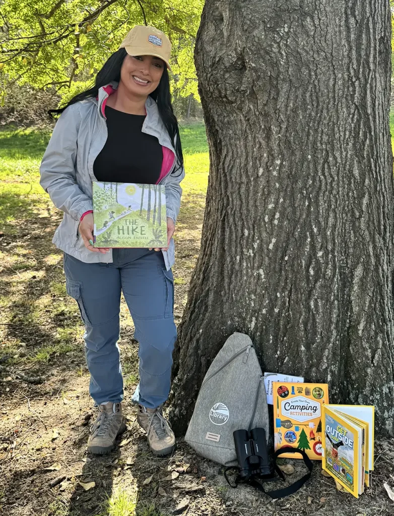 Library worker dressed for Spirit Week's Outdoors Day. She is wearing a hiking outfit and has checked out the Adventure Backpack.